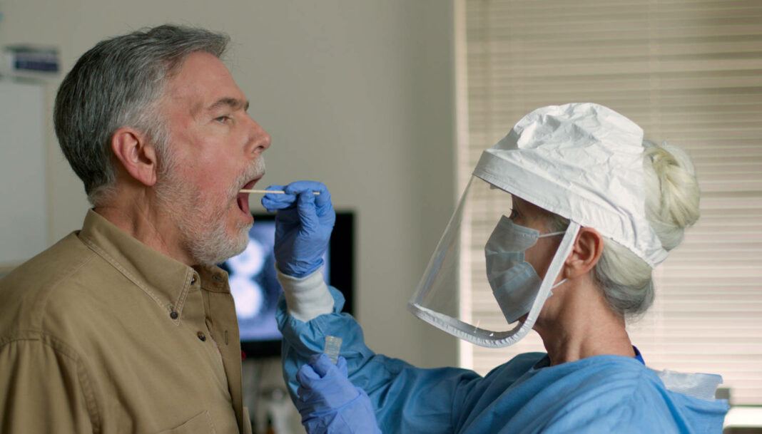 A mature Caucasian man in a clinical setting being swabbed by a healthcare worker